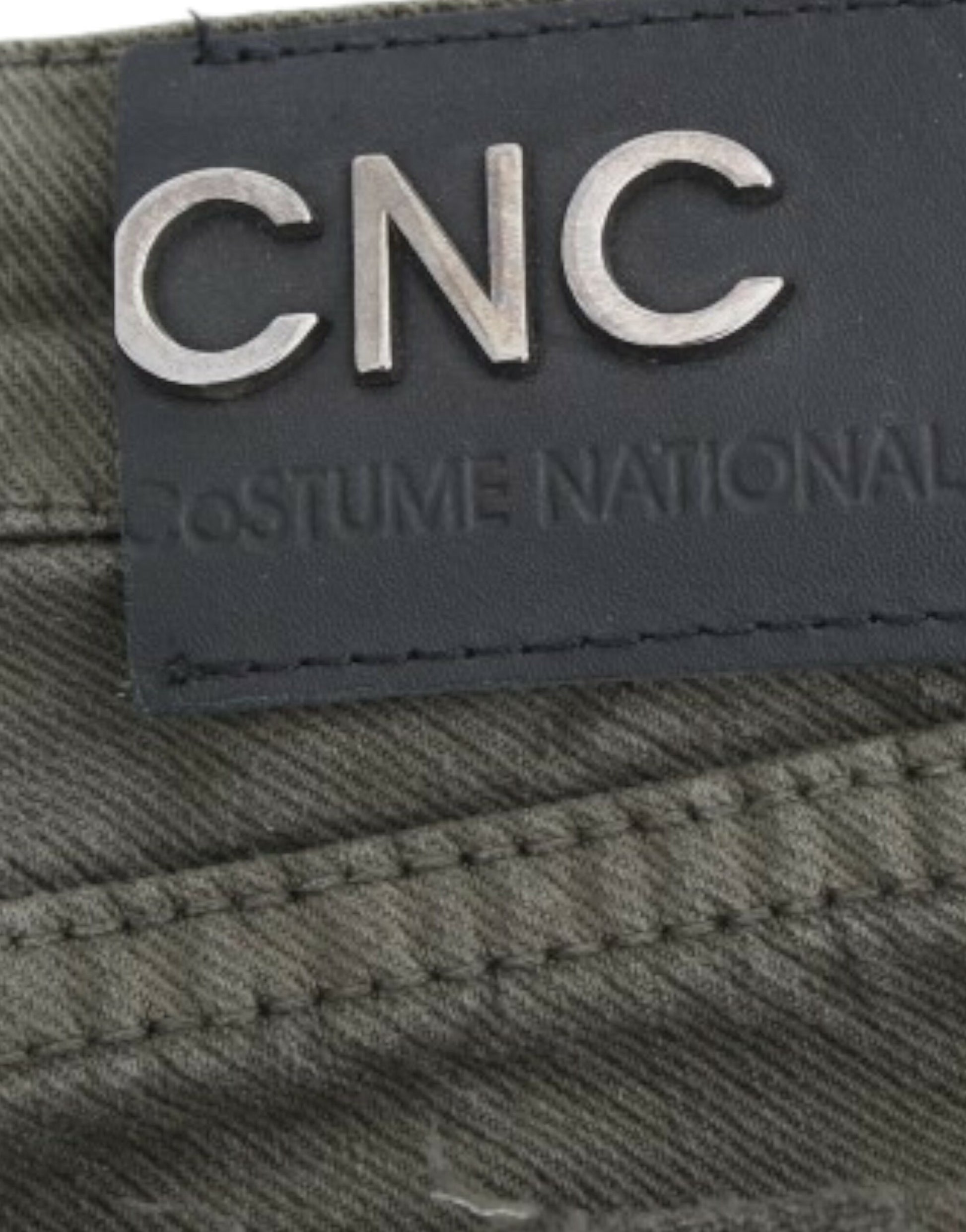 A close-up of a clothing label with the letters CNC prominently displayed in white on a black background. Below this, the