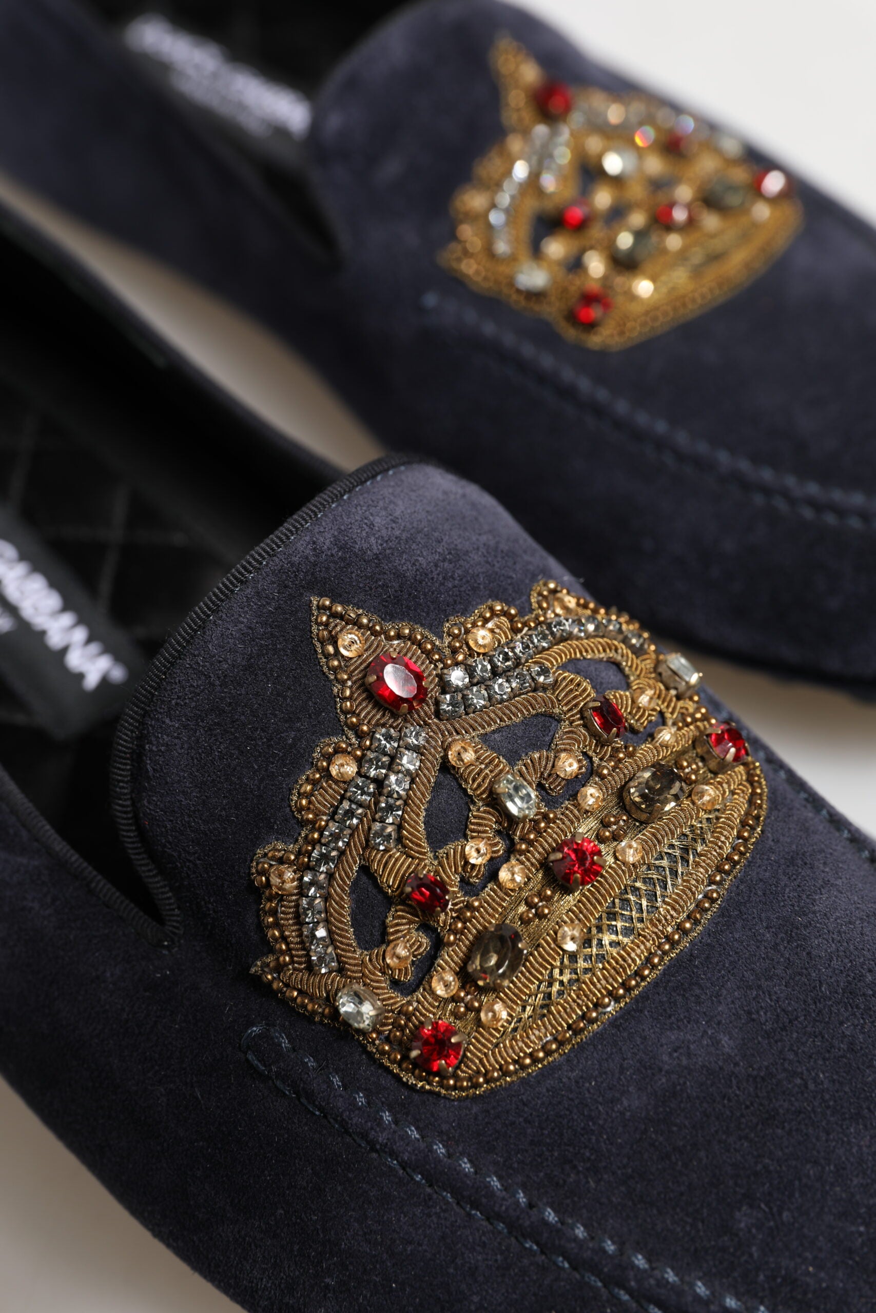 A pair of blue suede loafers with a crown embellishment featuring red and clear crystals